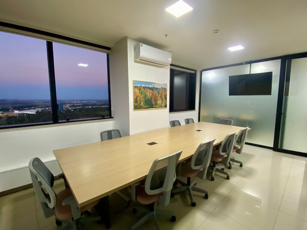 Sala de reuniões com mesa central retangular bege, quadro na parede, televisão e janela com a vista da cidade de Goiânia